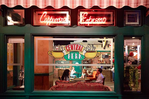 Exterior view of the Central Perk cafe with neon signs reading "Cappuccino" and "Espresso."
