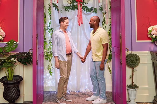 Two men holding hands and smiling in a set dressed for a wedding or ceremony.