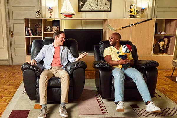Two men sitting in the black recliners on a recreation of Joey and Chandler's apartment set.