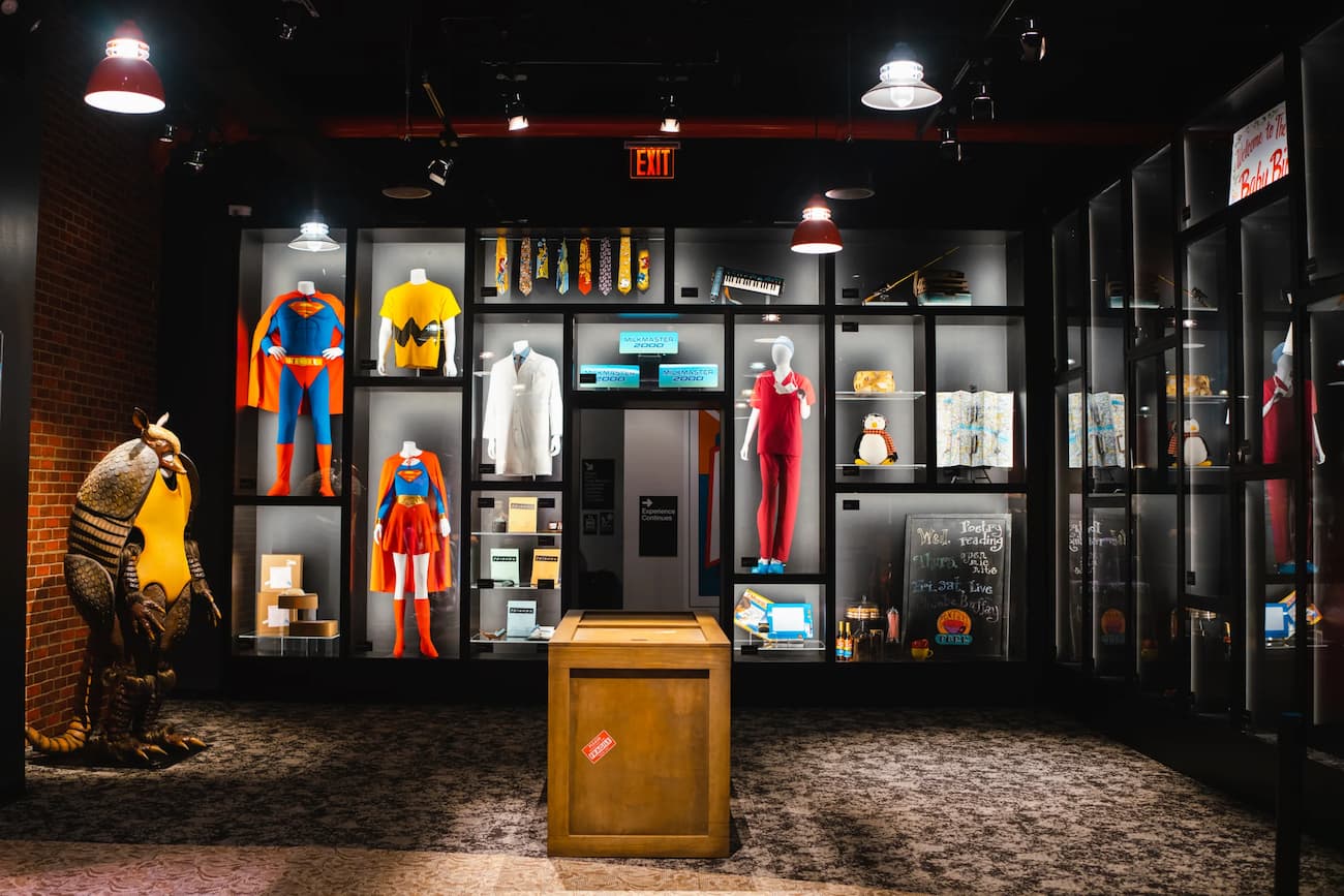 Wide view of a dark museum gallery with many FRIENDS costumes and props in lit cases.
